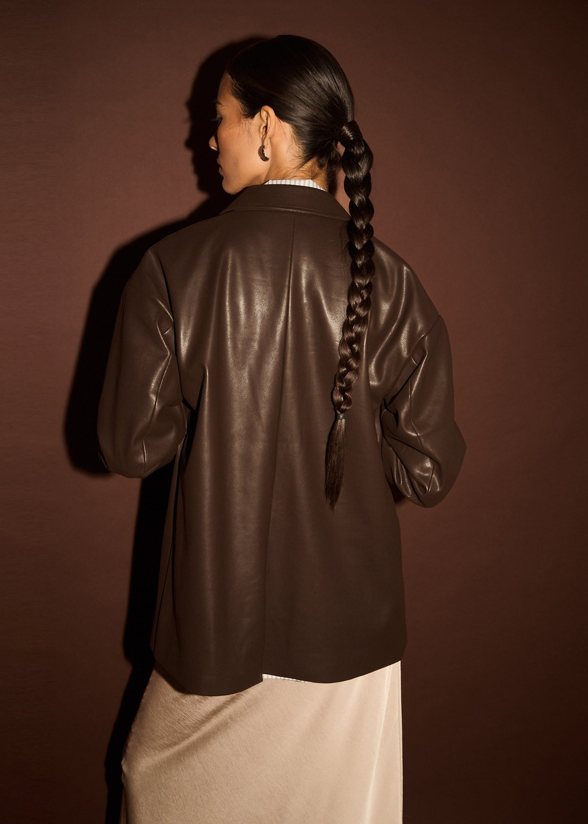 Close-up detail of the Chocolate Relaxed Vegan Leather Blazer, showing back shoulder, photographed against a brown backdrop.