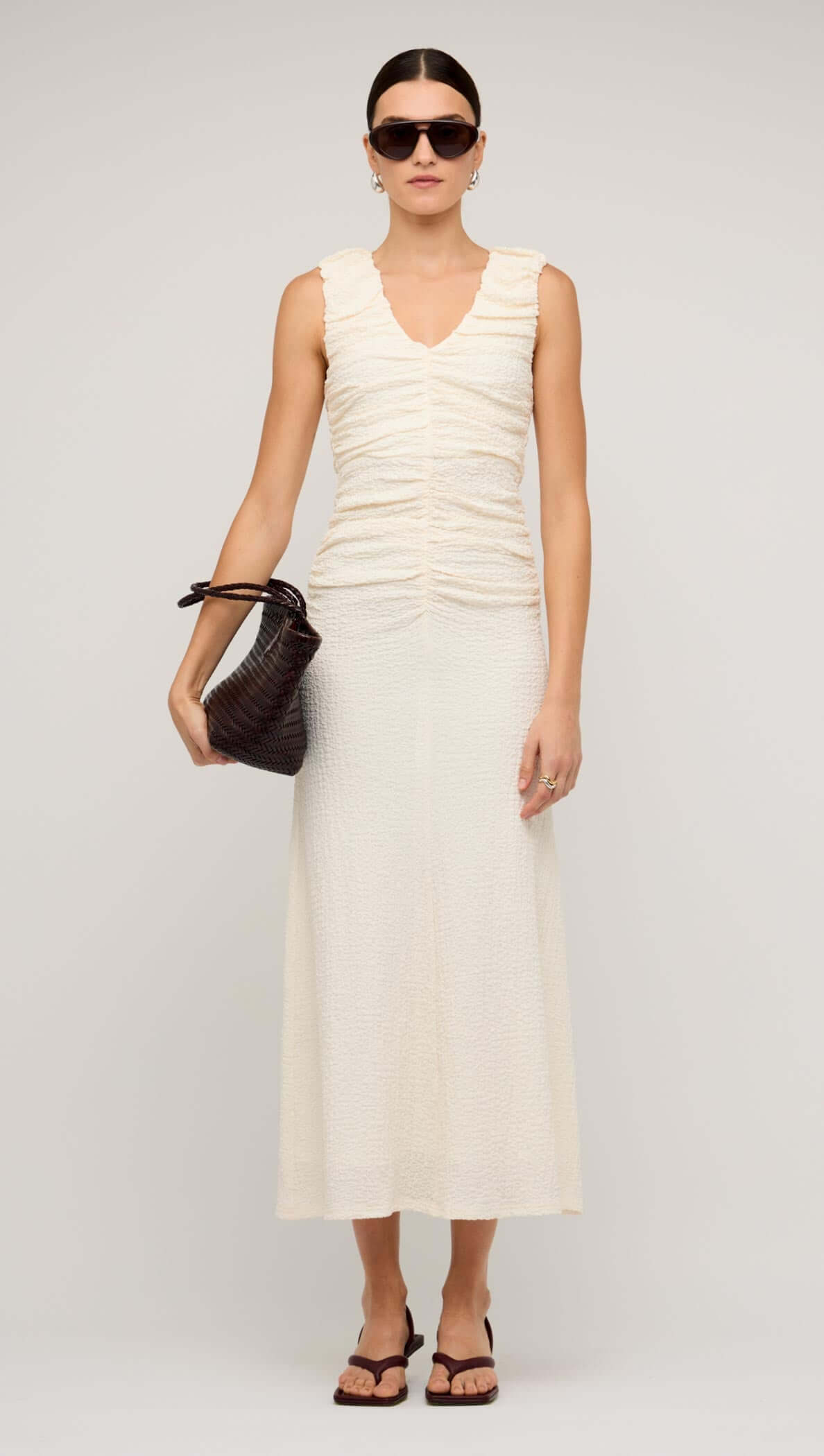 Woman wearing a cream gathered v-neck A-line midi dress, holding a handbag, and accessorized with sunglasses.