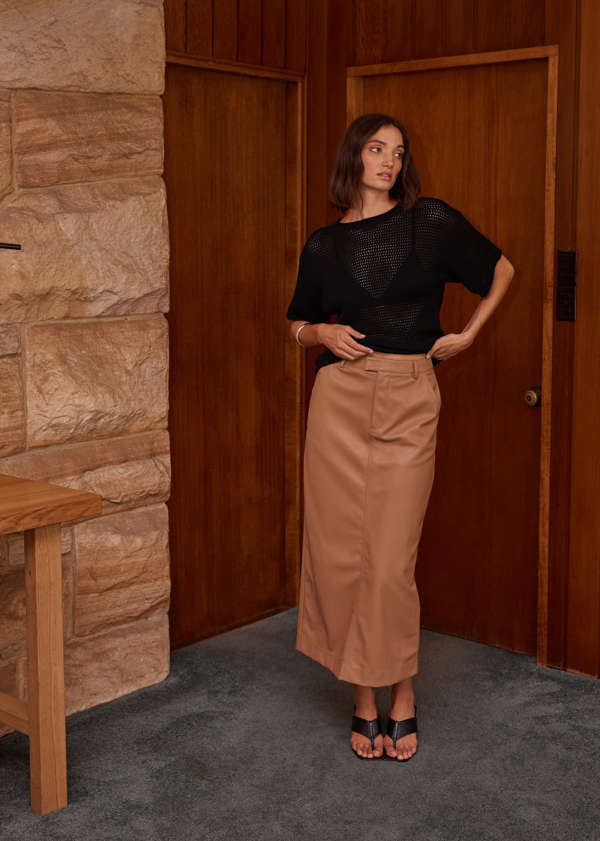 Model wearing Cynthia Pu Midi Skirt, showing belt loops, photographed against a timber doorway and stone wall.