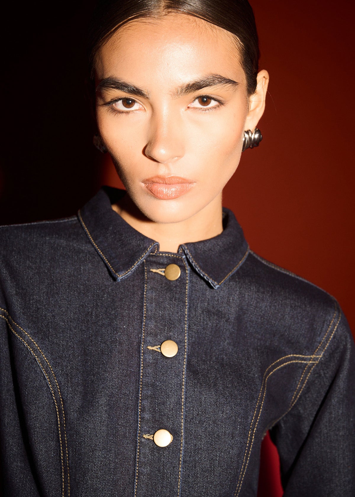Close-up detail of the Dark Wash Fitted Denim Shirt Jacket, showing collar and gold buttons, photographed against a red studio backdrop.