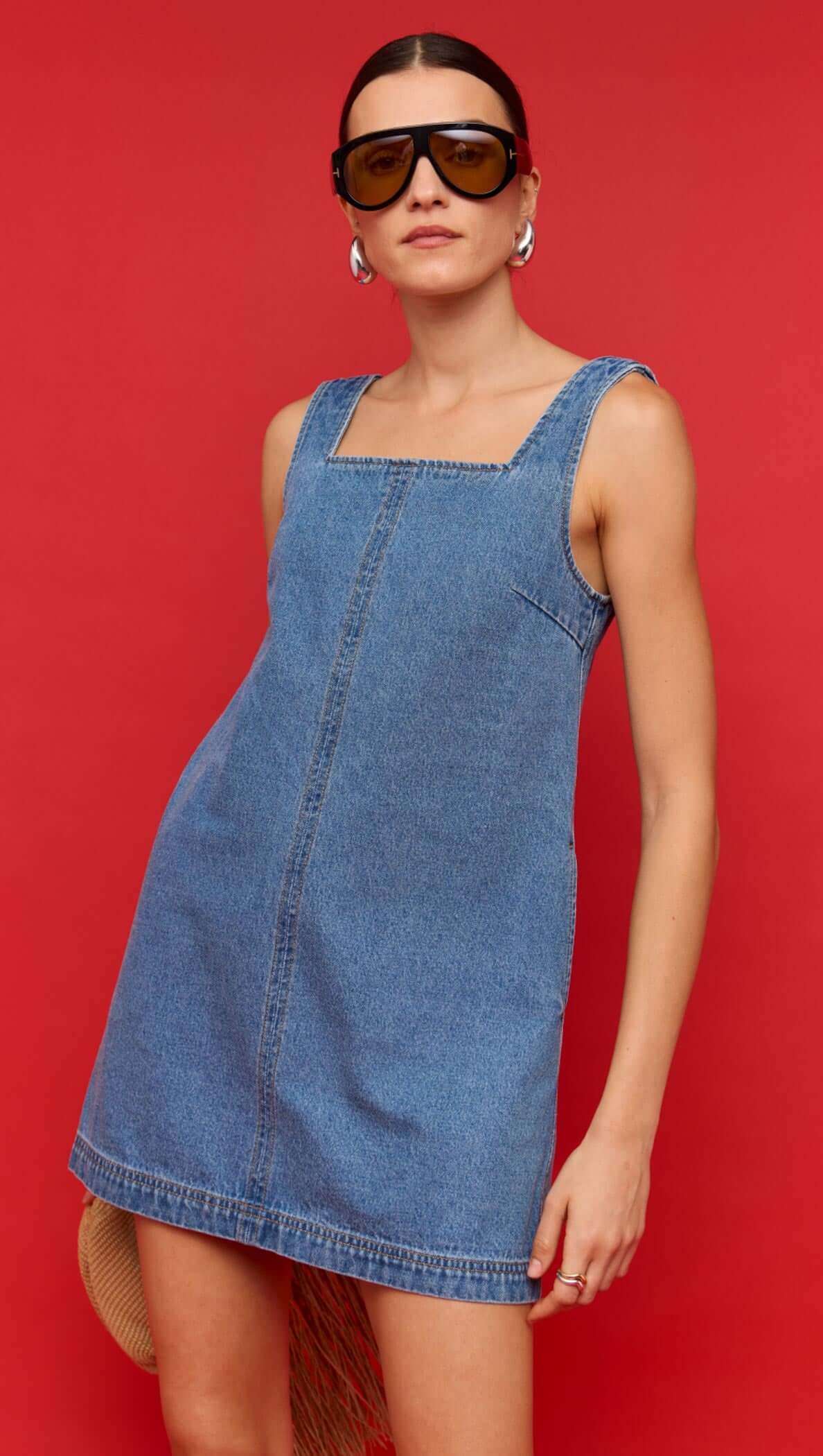 Denim Square Neck Mini Dress displayed on a model with a straw bag against a vibrant red background.