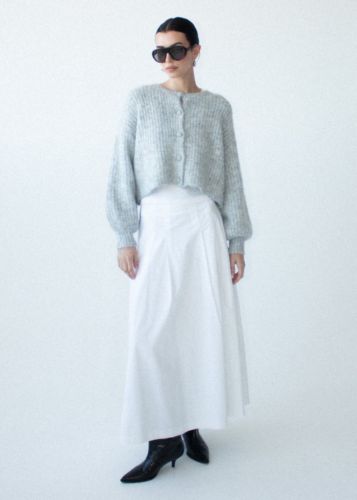 Model wearing Grey Marle Cropped Knit Cardigan, showing softly voluminous long sleeves, photographed against a pale blue studio.
