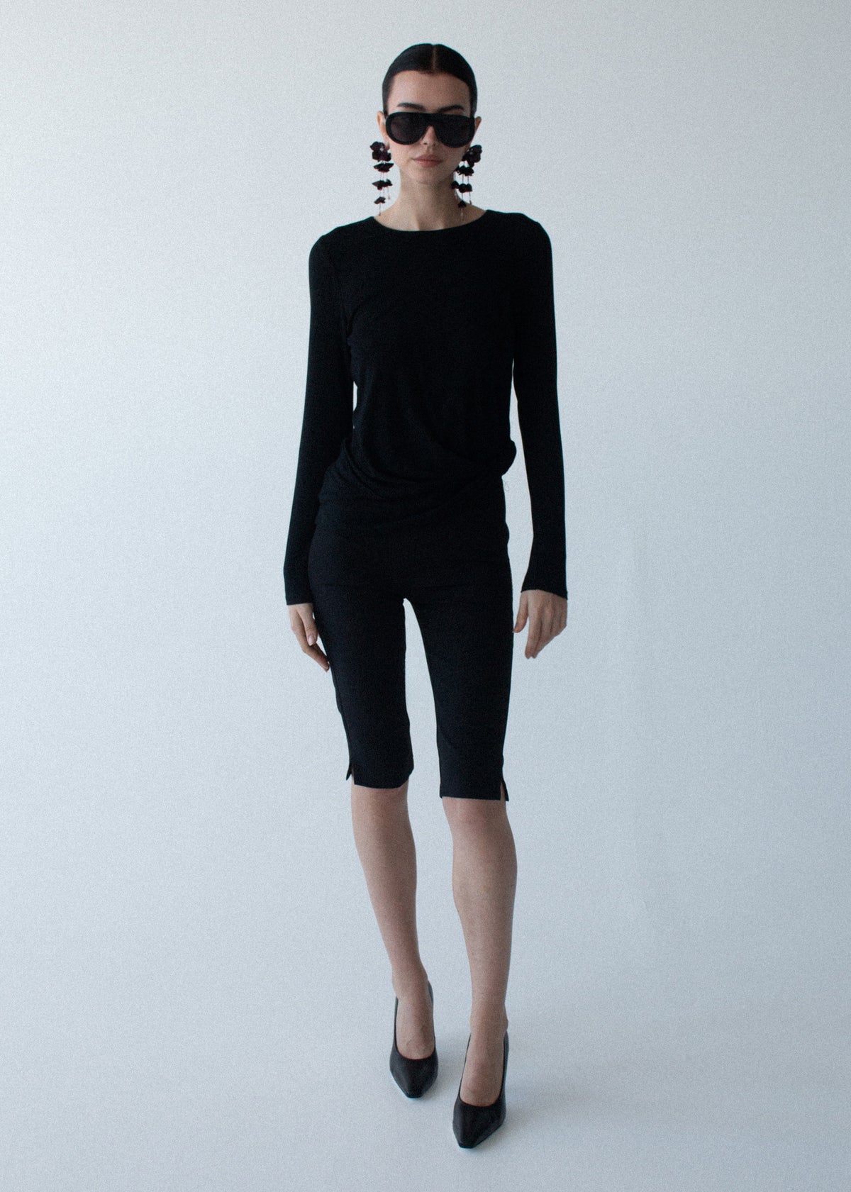 Model wearing Mid Rise Capri Pants in Black, showing a slim, streamlined silhouette and a capri length, photographed against a grey studio.