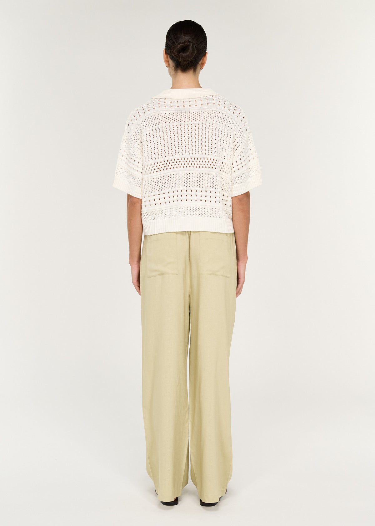 Back view of a woman wearing a Relaxed Crochet Polo Shirt in Cream with wide-leg pants, showcasing open crochet knit design.