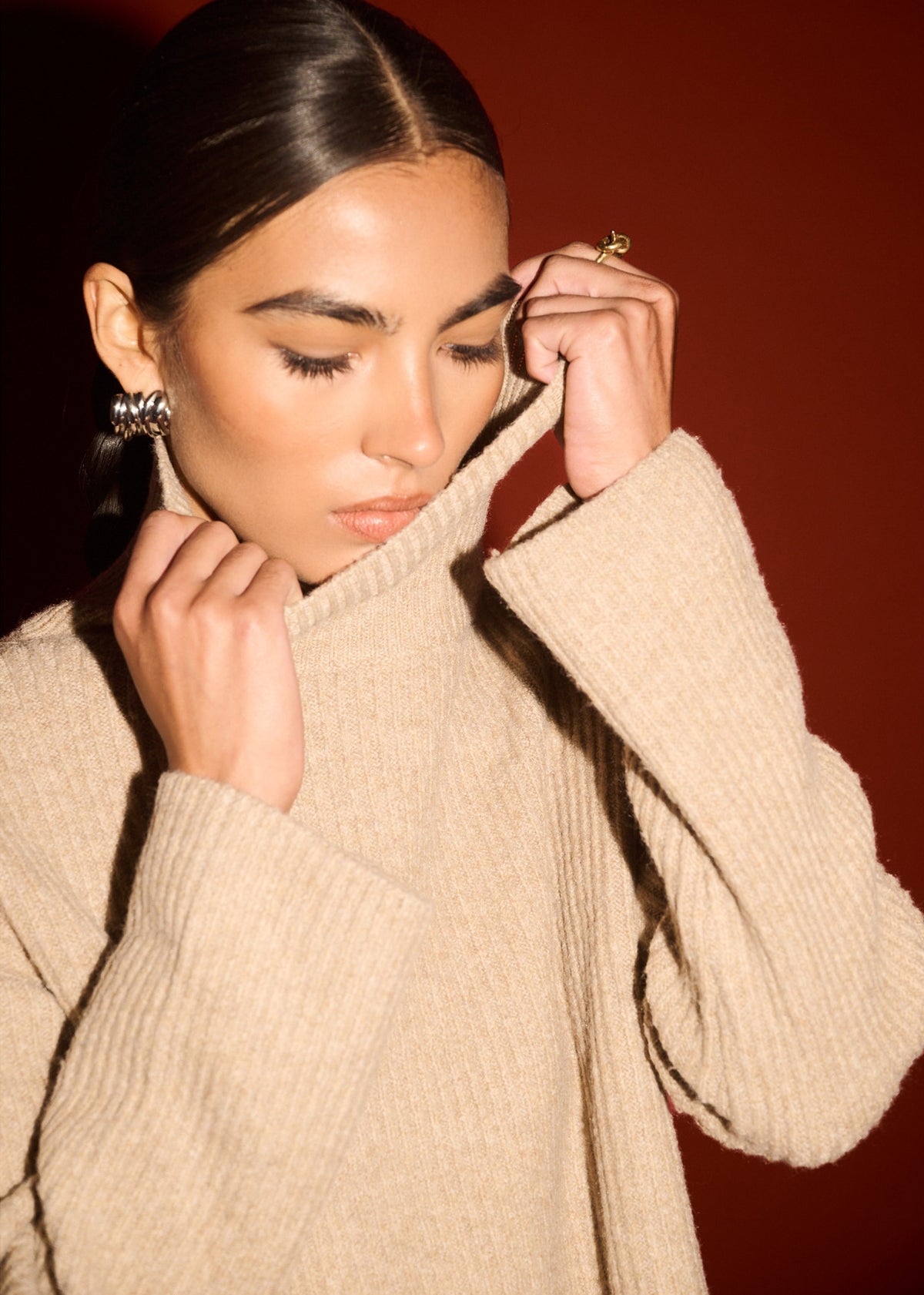 Close-up detail of the Oatmeal Roll Neck Oversized Knit Jumper, showing roll neck and sleeve, photographed against a red studio backdrop.