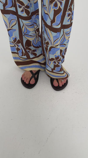 Model wearing patterned pants with blue and brown floral design and black flip-flops, showcasing a casual summer look.