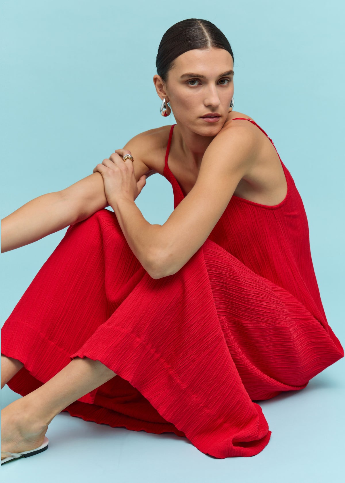 Model seated wearing Red Crepe V-Neck Floaty Maxi Dress with slim adjustable straps, photographed against a turquoise blue backdrop.
