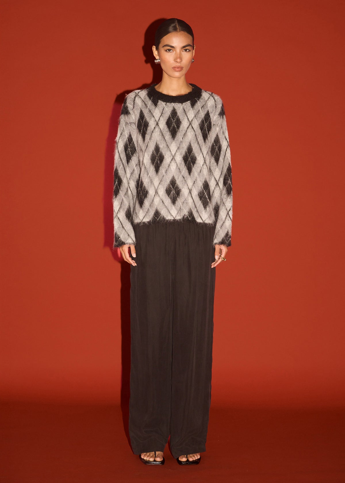 Model wearing Relaxed Fit Argyle Brushed Jumper with a relaxed fit and a crew neckline, photographed against a rust red backdrop.