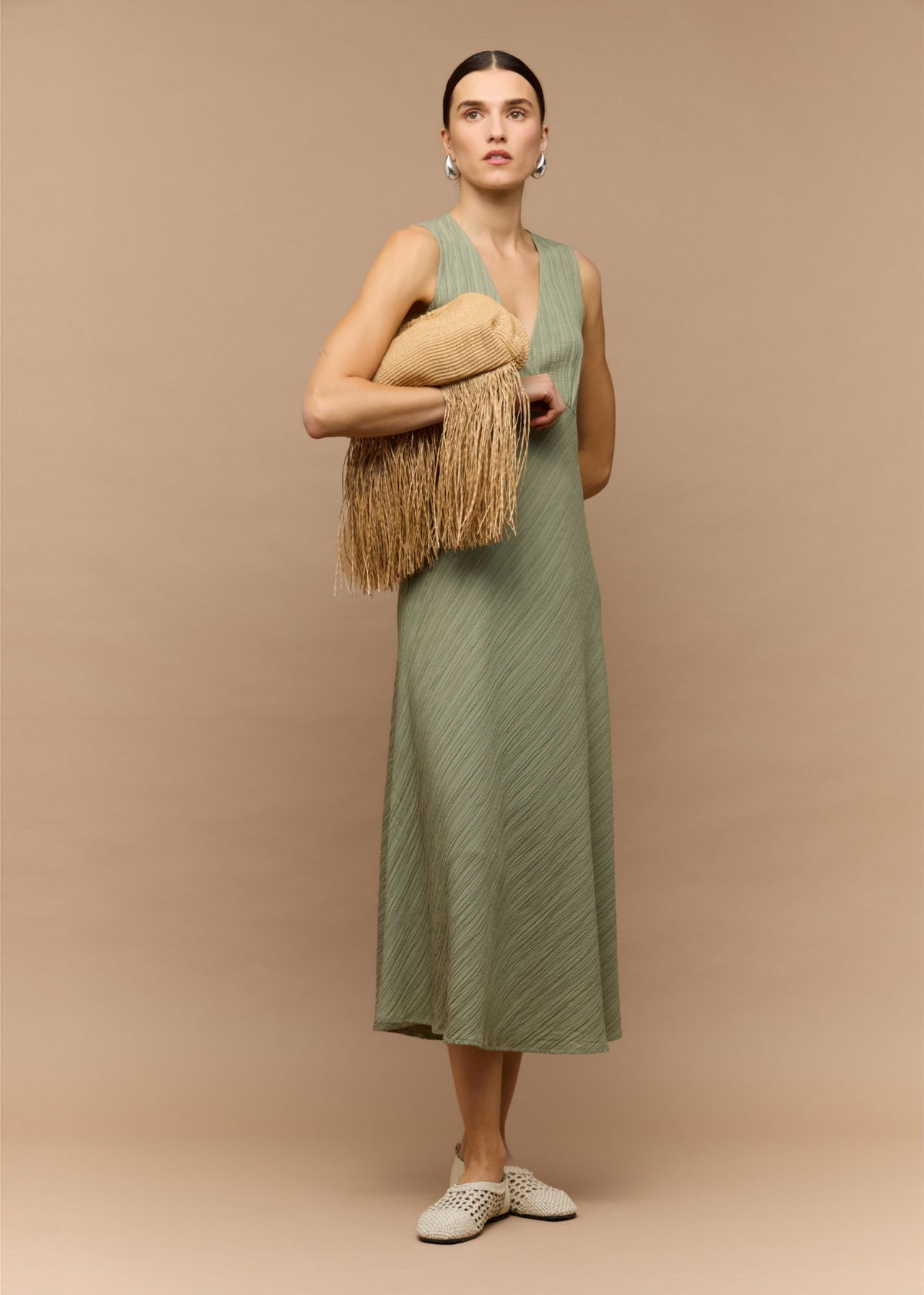 Model wearing Sage Green V-Neck Bias Midi Dress with a sage green colour-way and a sleeveless cut, turned to the side.