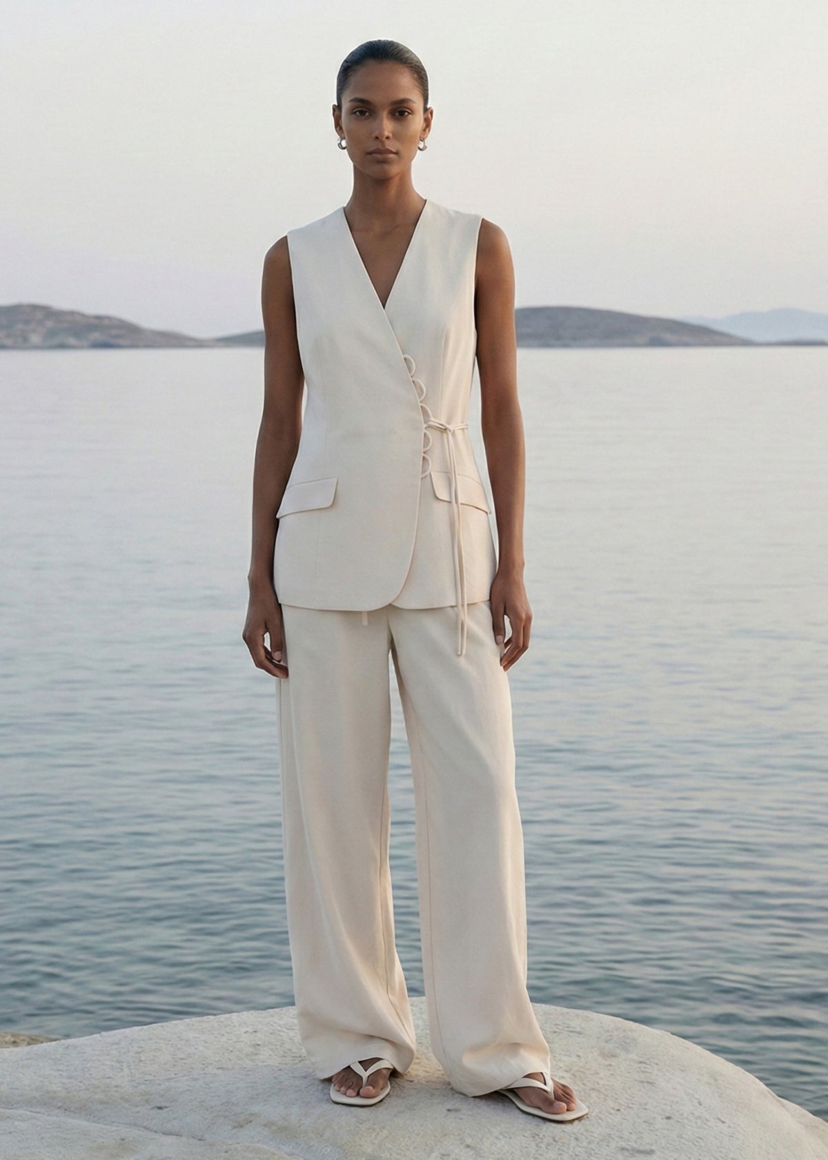 Model wearing Stone Cotton Wrap Front Vest, showing a scalloped loop-and-tie fastening and flap pockets, photographed on rocks by the sea.