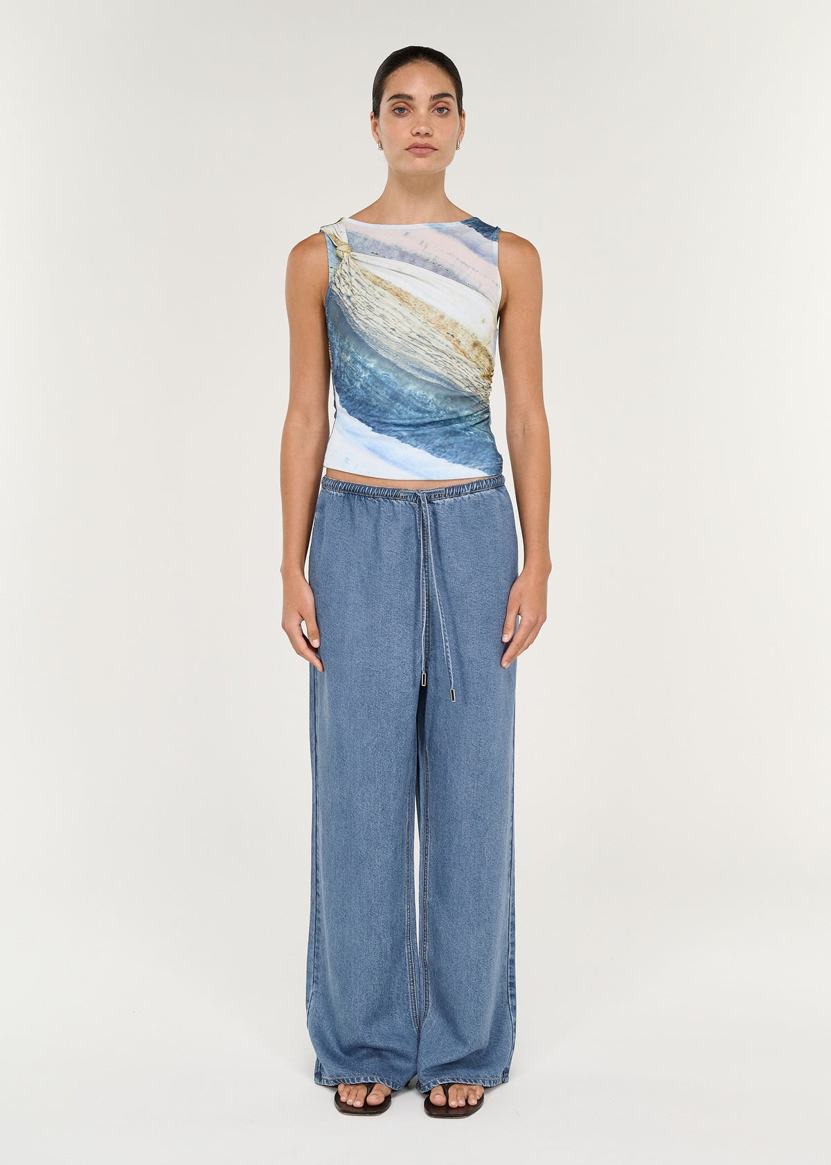 Model wearing Sleeveless Mesh Twist Top in Ocean Tide paired with relaxed denim pants, showcasing coastal-inspired print and twisted detail.